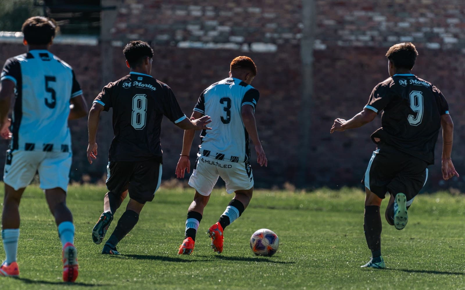 Youth soccer players in numbered team kits chasing the ball across a grass field