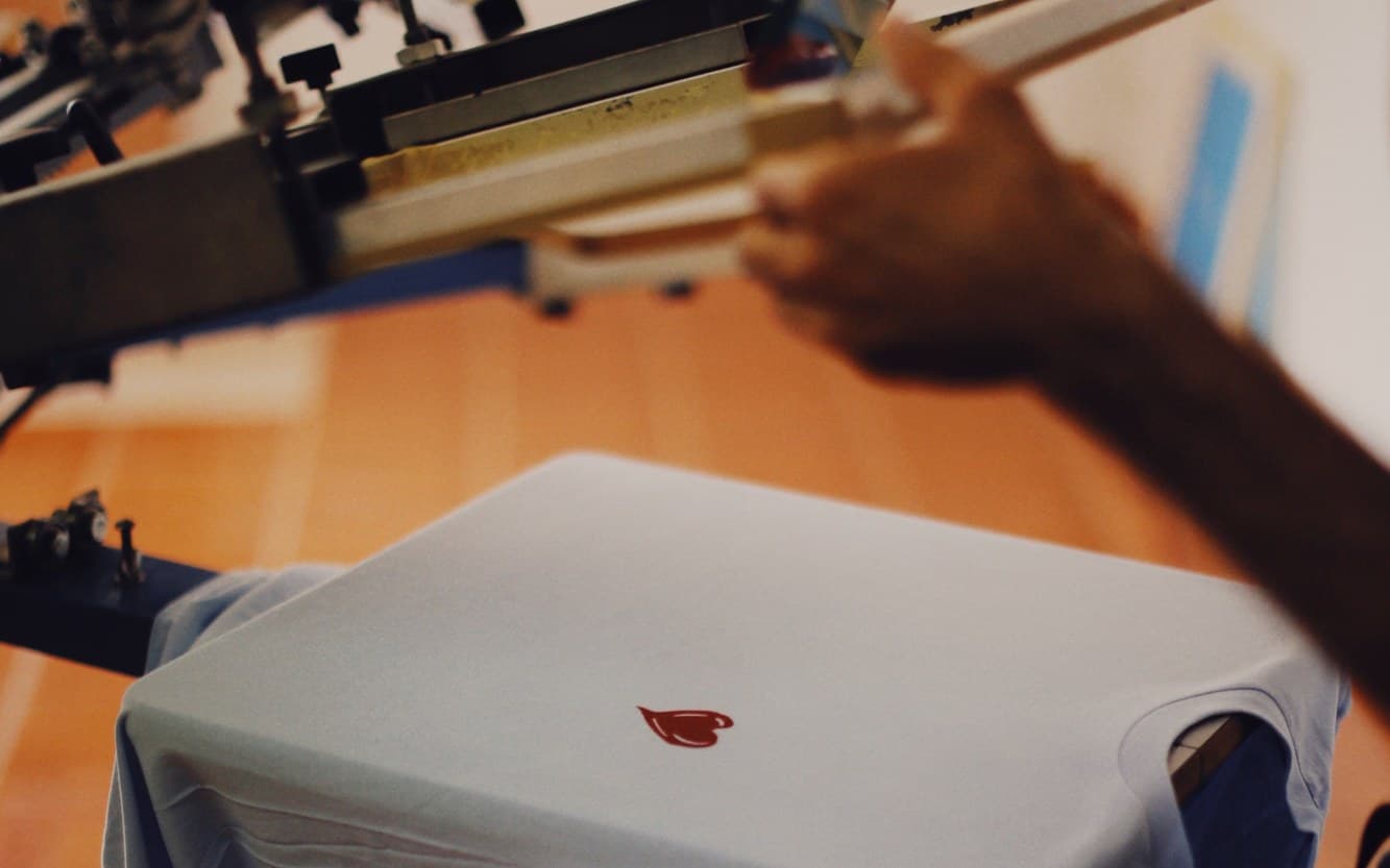 A printer lowering a screen printing press over a light blue tee with a small red heart printed on the chest