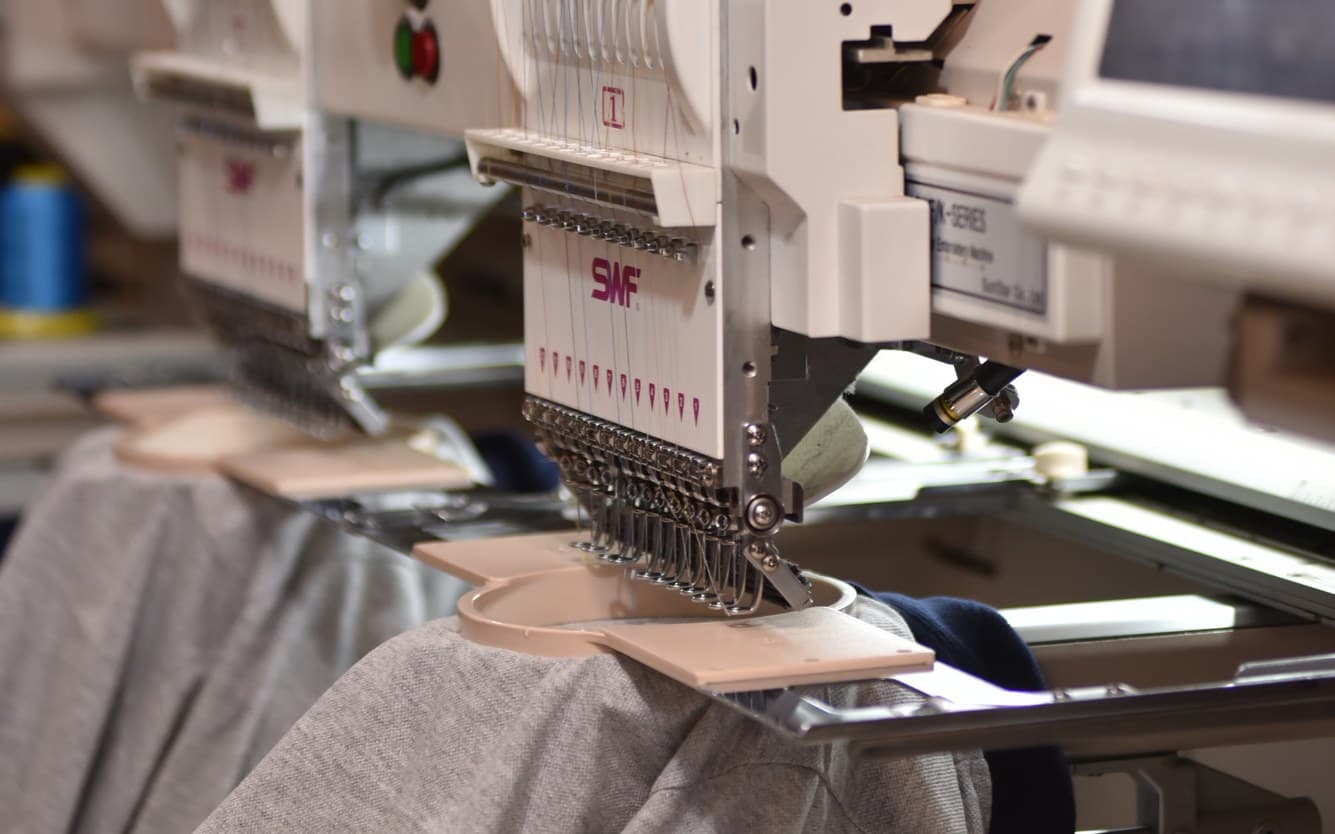 A row of multi-head embroidery machines with gray shirts hooped under the needles, spools of colored thread on the shelves behind