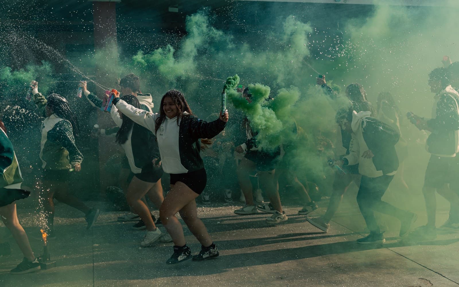 Students in matching green and white school hoodies running through green smoke and confetti at a spirit rally